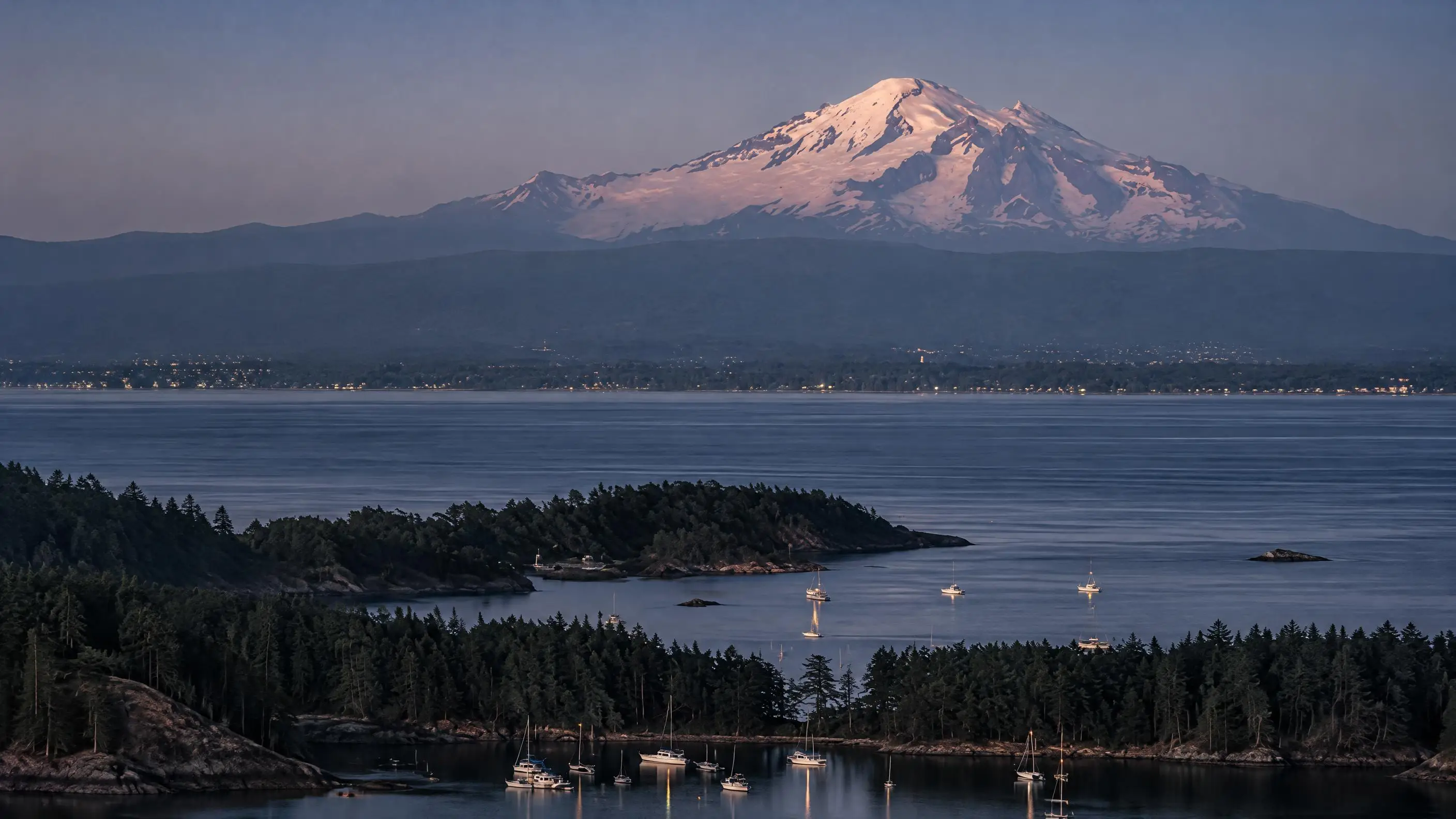 Mount Baker above Bellingham Bay at dusk, sailboats at anchor in a wooded cove