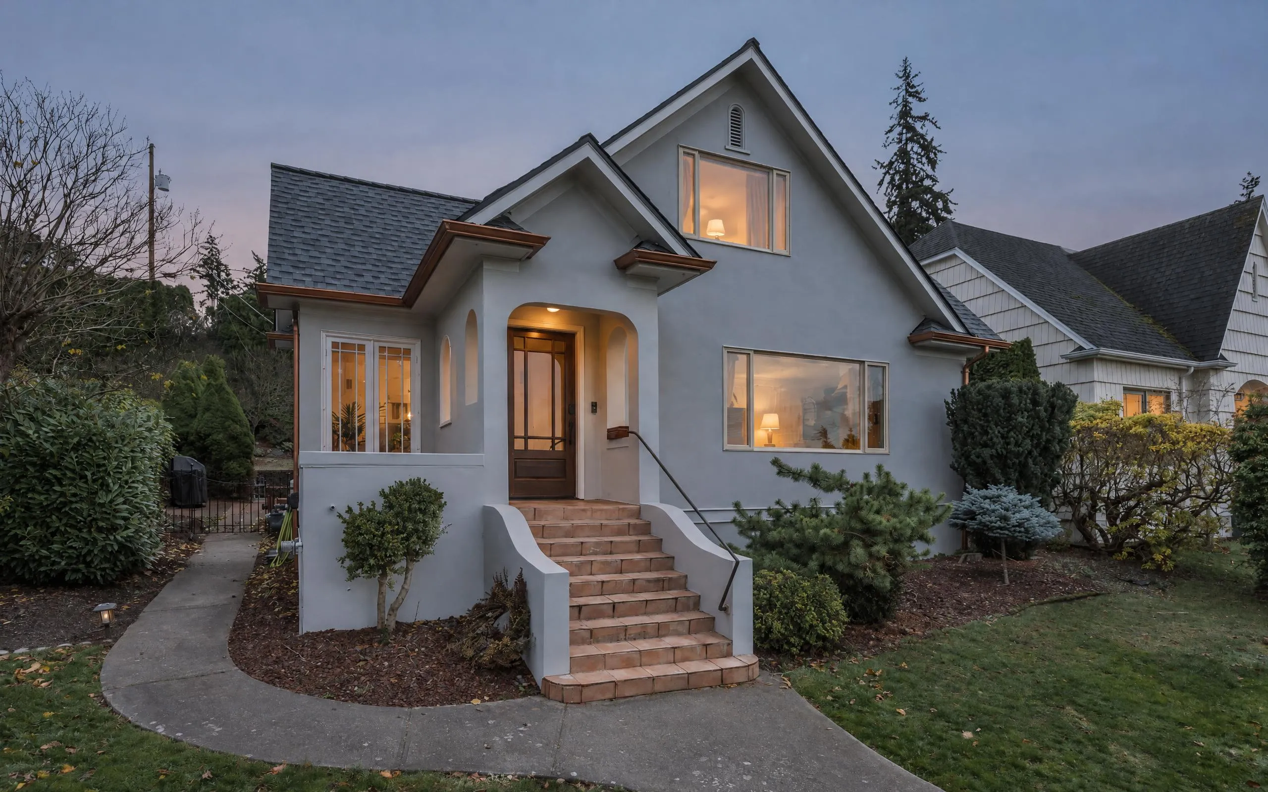 An older stucco house with a steep gabled roof and tiled front steps, at dusk with the lights on